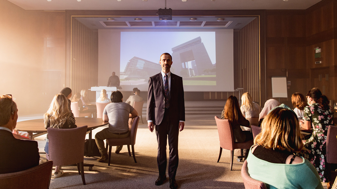 The Dean of London Business School stands facing the camera in a meeting room full of students