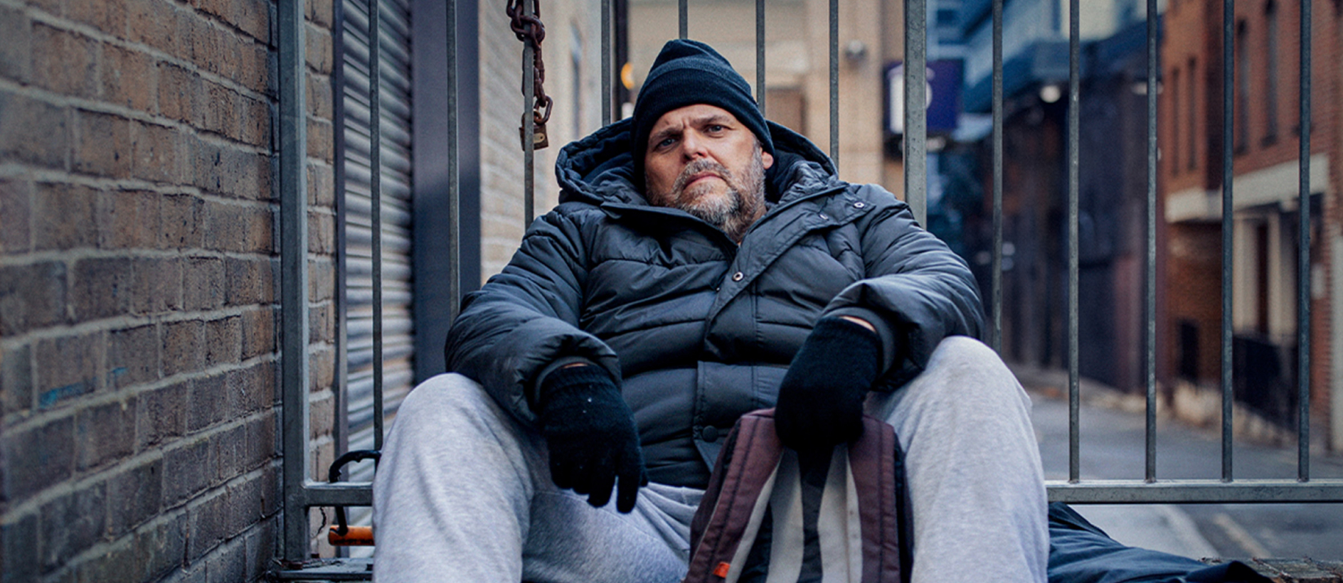 Homeless man in winter clothes sitting on the ground against metal railings
