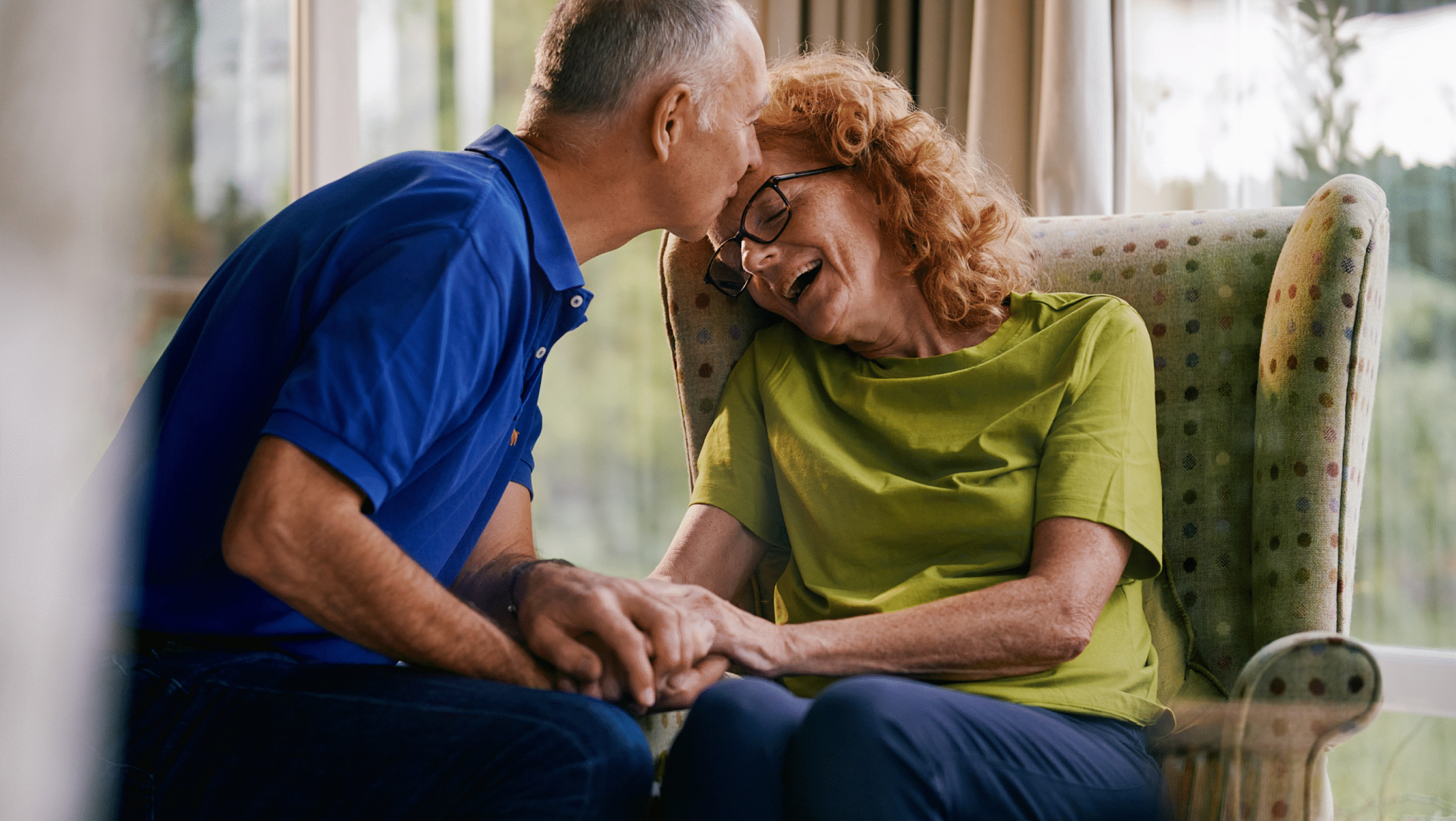 A woman with MND sits in an arm chair, her partner kisses her forehead.