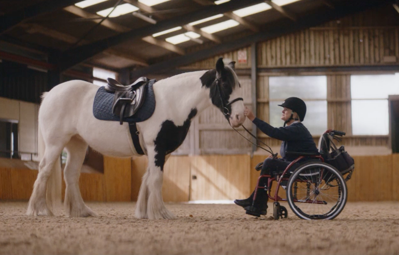 Woman in a wheelchair strokes the nose of a black and white horse wearing a saddle